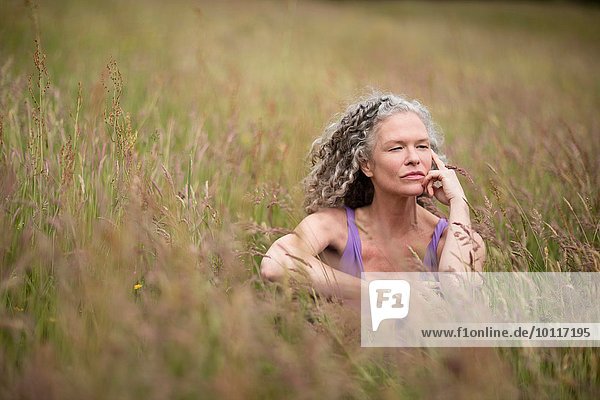 Reife Frau sitzt im langen Gras und schaut weg.