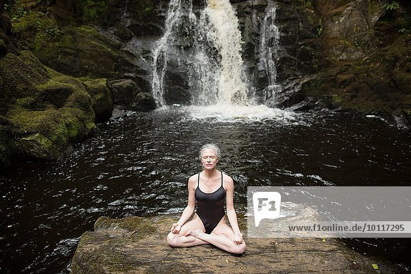 Reife Frau übt Lotus-Pose vor dem Wasserfall