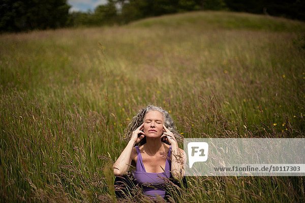 Reife Frau mit Fingern an den Schläfen  die in langem Gras sitzt und meditiert.