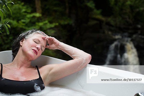 Reife Frau mit geschlossenen Augen im Schaumbad vor dem Wasserfall beim Öko-Retreat