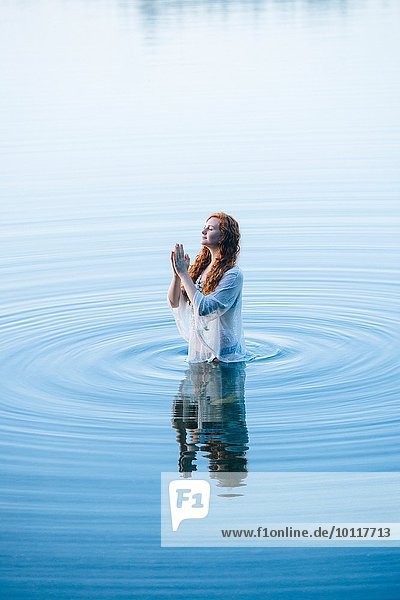 Junge Frau steht mit geschlossenen Augen und zusammengehaltenen Händen im See.