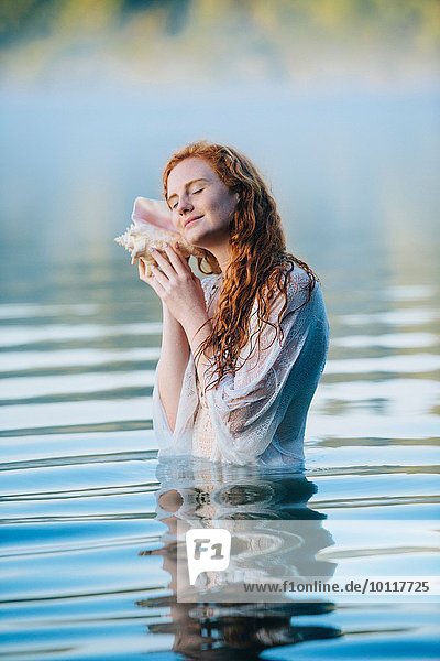 Young woman standing in misty lake with eyes closed listening to seashell