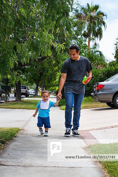 Young man holding hands with toddler brother whilst strolling along suburban sidewalk