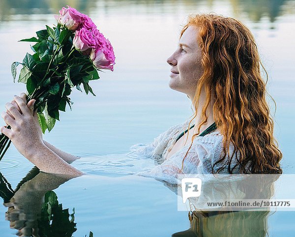 Kopf und Schultern einer jungen Frau mit langen roten Haaren im See,  die auf rosa Rosen blickt.