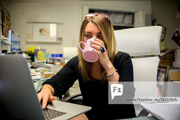 Young woman in office  sitting at desk  drinking coffee  using laptop