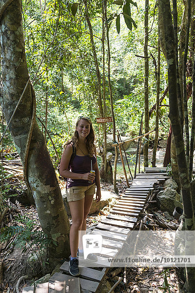 Tourist smiling  holding a drink  standing on a boardwalk in the woods  path to the Na Muang waterfalls  Ko Samui  Thailand  Asia