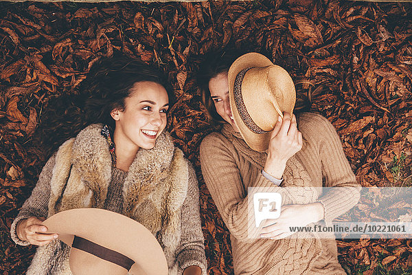 Two smiling female friends lying side by side on autumn leaves in an park