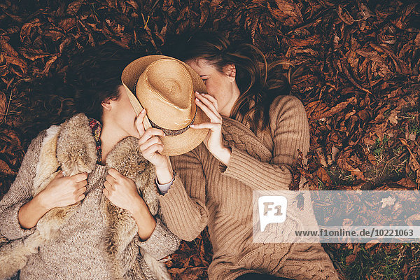 Two female friends lying side by side on autumn leaves in an park hyding behind a hat
