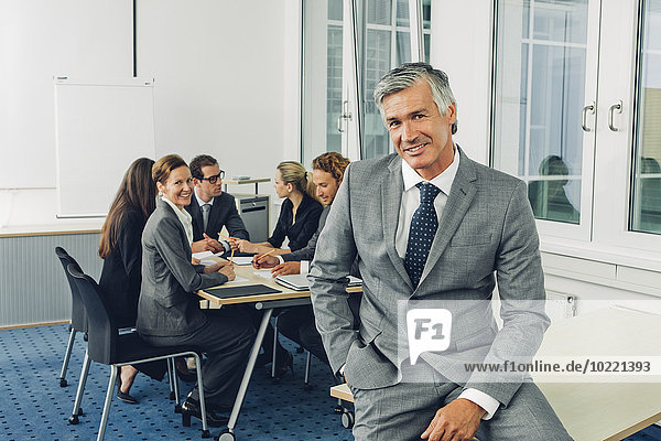 Mature businessman sitting on desk  team working in background