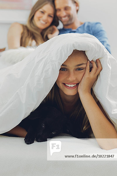 Porträt eines glücklichen Mädchens mit Welpe im Bett und Eltern im Hintergrund