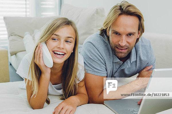 Father and daughter lying on couch using laptop and telephone