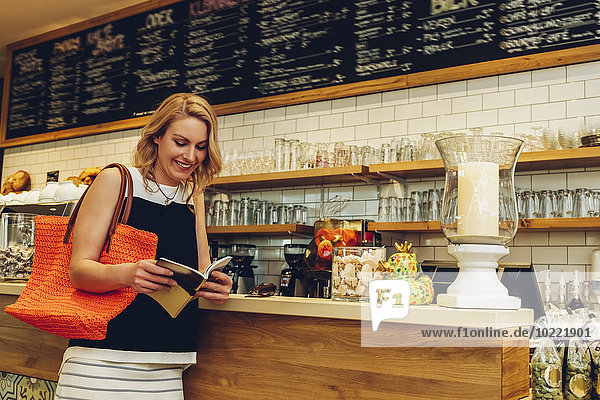 Lächelnde blonde Frau steht an der Theke in einem Coffee Shop und schaut auf ein Booklet