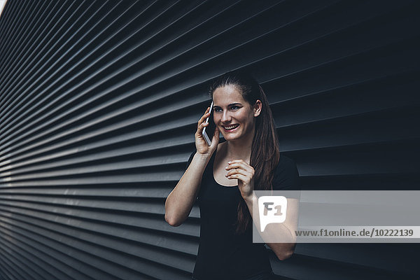 Porträt einer lächelnden jungen Frau beim Telefonieren mit Phablet vor schwarzer Fassade