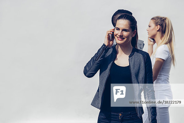 Porträt einer jungen Frau beim Telefonieren mit dem Smartphone vor zwei weiteren Personen