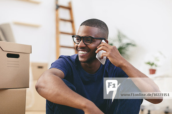 Porträt eines glücklichen jungen Mannes  der neben Kartons sitzt und mit dem Smartphone telefoniert.