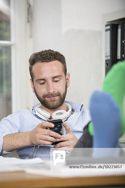 Junger Mann im Büro mit Füßen auf dem Schreibtisch und Blick aufs Handy