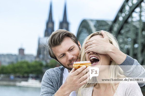Deutschland  Köln  junge Frau  die einen Bagel probiert  während ihr Freund ihr die Augen zudeckt