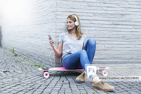 Deutschland  Köln  junge Frau sitzt auf rosa Skateboard und hört Musik mit Kopfhörern.
