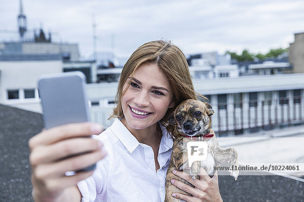 Porträt einer lächelnden jungen Frau  die einen Selfie mit ihrem Hund auf der Dachterrasse nimmt.