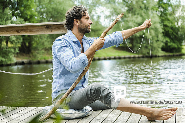 Mann sitzt auf der Plattform am Wasser mit selbstgemachter Angelrute