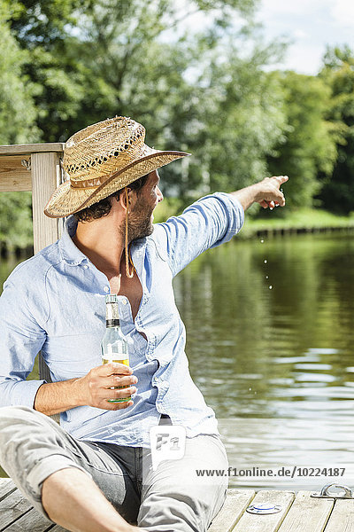 Mann sitzt auf der Plattform am Wasser mit einer Bierflasche  die mit dem Finger auf ihn zeigt.