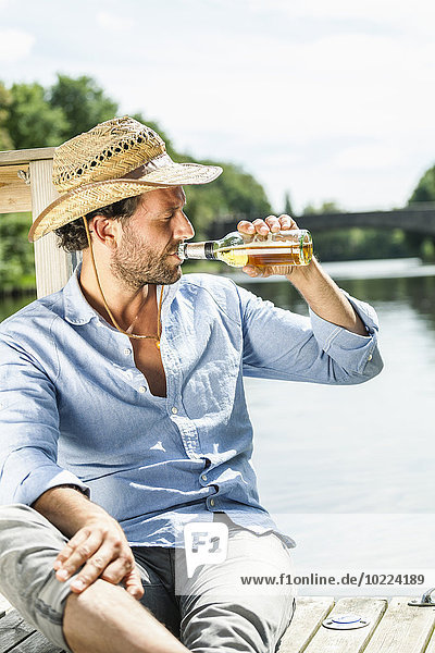 Mann sitzt auf der Plattform am Wasser und trinkt Bier.