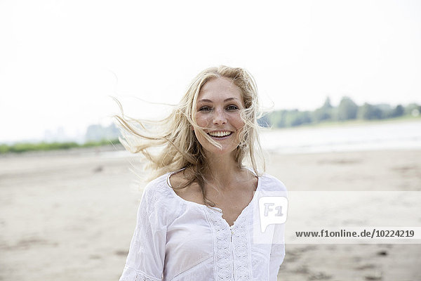 Porträt einer lächelnden blonden Frau mit blasendem Hait am Strand