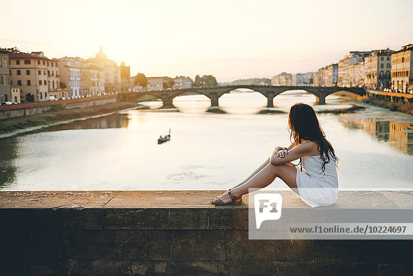 Italien Florenz Frau in weißem Sommerkleid bei Sonnenuntergang auf einer Brücke sitzend