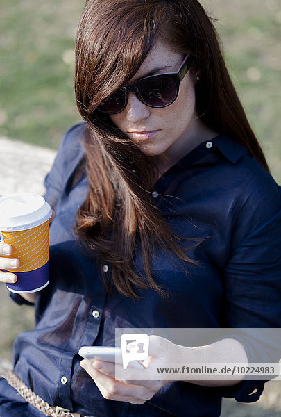 Porträt einer Frau mit Sonnenbrille auf einer Parkbank mit Smartphone und Kaffee zum Mitnehmen