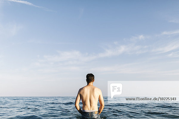 Rückansicht eines jungen Mannes,  der im Meer steht und in die Ferne schaut.