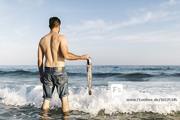 Back view of young man standing in the water of the sea holding caught fish