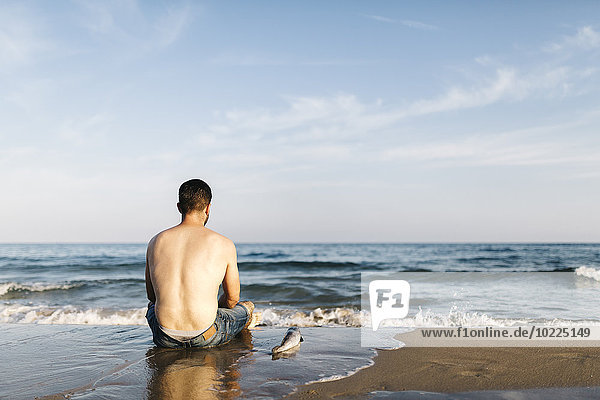 Back view of young man sitting at seafront beside a caught fish