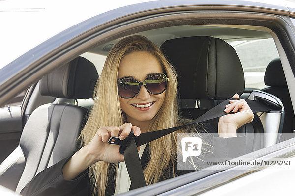 Portrait of smiling blond woman with sunglasses putting on safety belt in her car