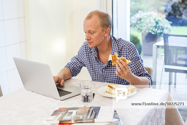 Mann zu Hause mit seinem Laptop beim Essen eines Snacks