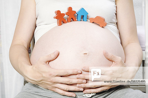Schwangere Frau vor dem Kinderbett mit Spielzeughaus  Spielfiguren und Spielzeugauto auf dem Bauch