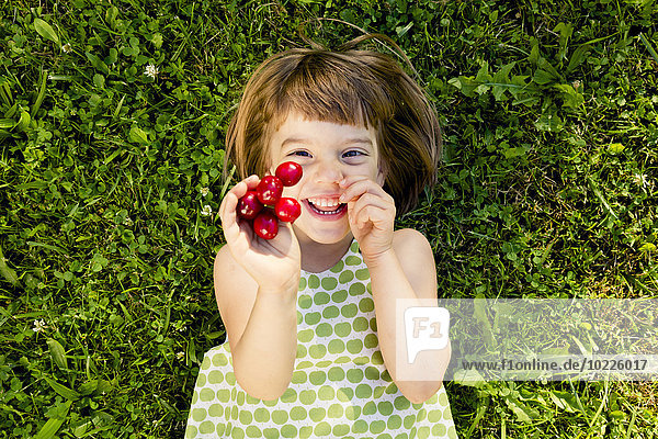 Lächelndes kleines Mädchen mit Kirschen auf einer Wiese liegend