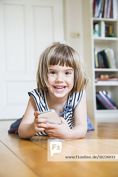 Portrait eines glücklichen kleinen Mädchens auf Holzboden liegend mit Smartphone