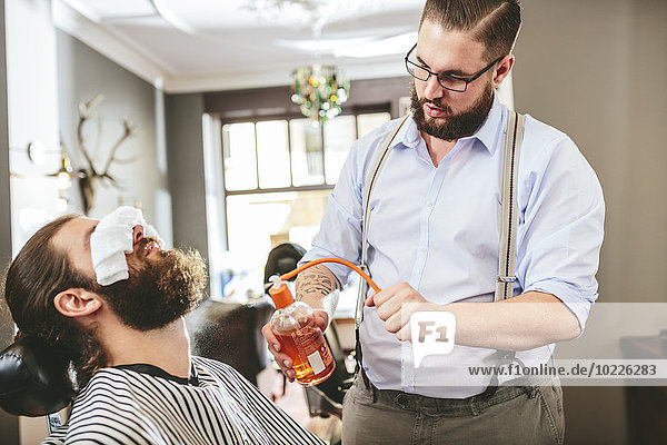 Barber spraying aftershave on beard of a customer