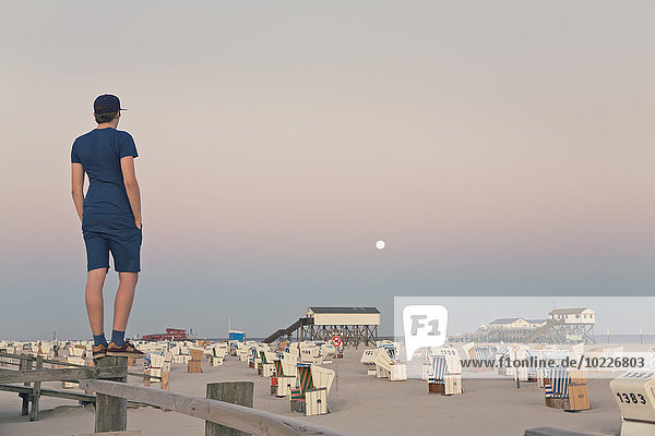 Deutschland  St. Peter-Ording  junger Mann auf Holzzaun stehend mit Blick auf den Strand