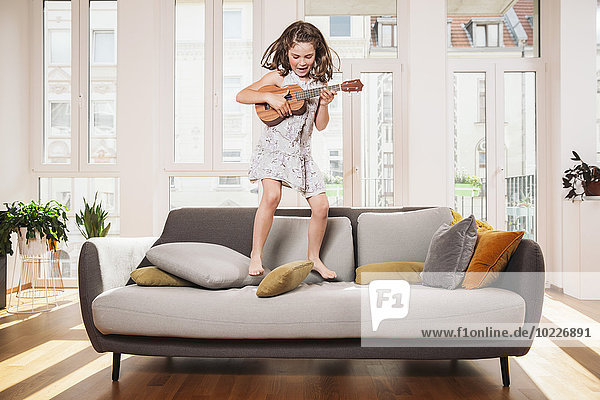 Glückliches Mädchen spielt Minigitarre beim Springen auf einer Couch im Wohnzimmer zu Hause.