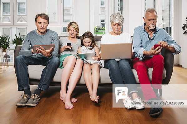 Gruppenbild von drei Generationen Familie  die auf einer Couch mit verschiedenen digitalen Geräten sitzt.