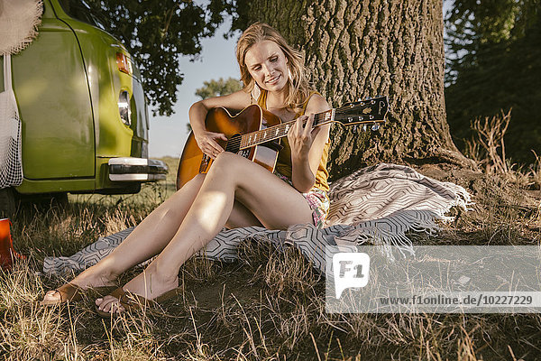Young woman playing guitar at tree beside van