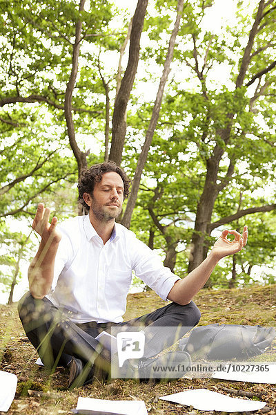 Germany  businessman in forest surrounded by papers practising yoga