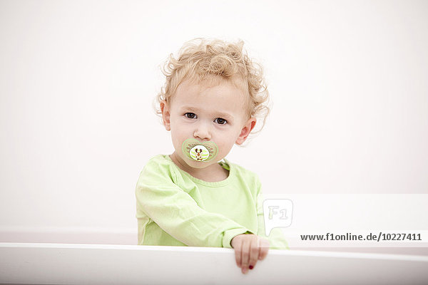 Porträt des kleinen blonden Mädchens mit Schnuller im Kinderbett stehend