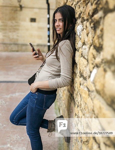 Young woman with smartphone leaning against stone wall