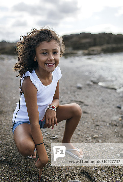 Spanien  Gijon  Porträt eines lächelnden kleinen Mädchens  das am Strand kauert