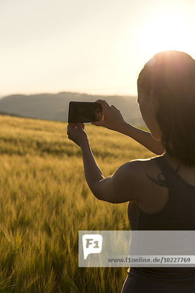 Deutschland  Frau  die bei Sonnenaufgang vor einem Feld steht und mit ihrem Smartphone fotografiert.