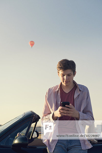 Teenager with smartphone leaning at convertible car