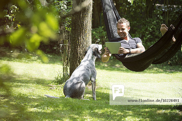 Entspannter Mann in der Hängematte liegend mit digitalem Tablett und Hund neben ihm
