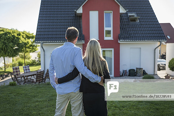 Ein glückliches Paar steht Arm in Arm im Garten und schaut auf sein Haus.
